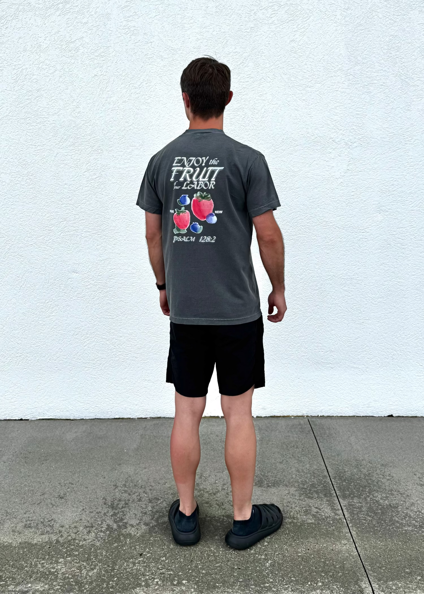 Man wearing a gray t-shirt with fruit design and text, standing against a white wall.