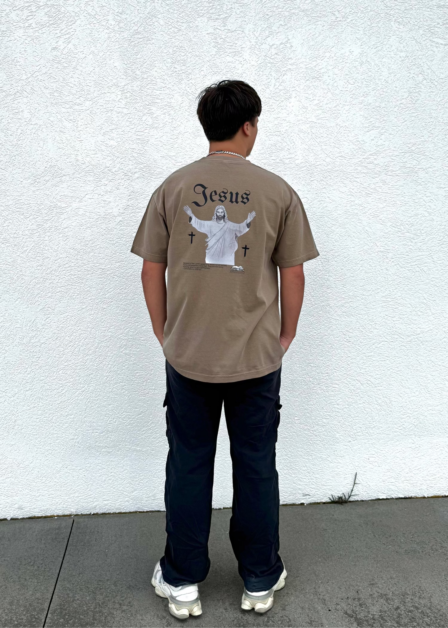 Person wearing a beige t-shirt with 'Jesus' printed on the back, standing against a white wall.