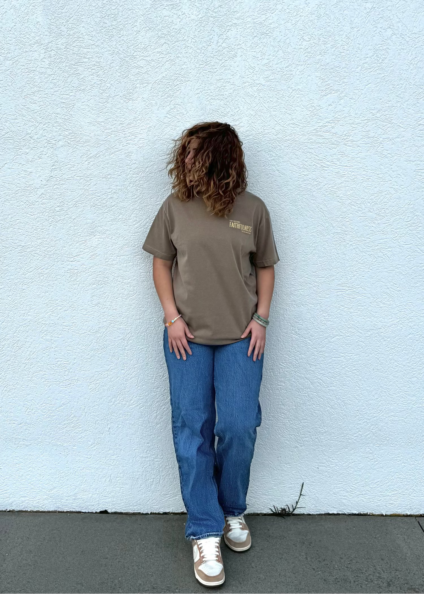 Person wearing a brown t-shirt and blue jeans standing against a light gray wall.