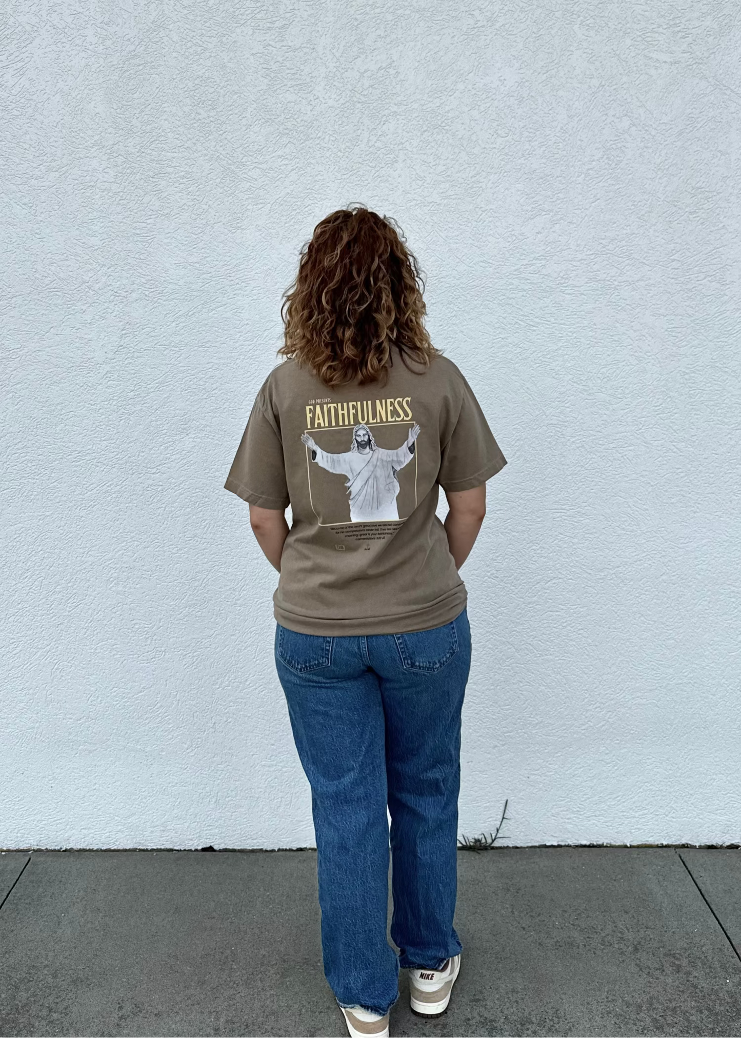 Person wearing a brown t-shirt with a graphic and text, standing against a light gray wall.
