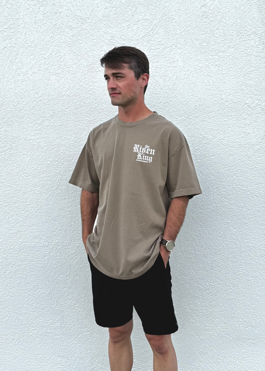 Man wearing a brown 'Risen King' t-shirt with text and black shorts against a white wall