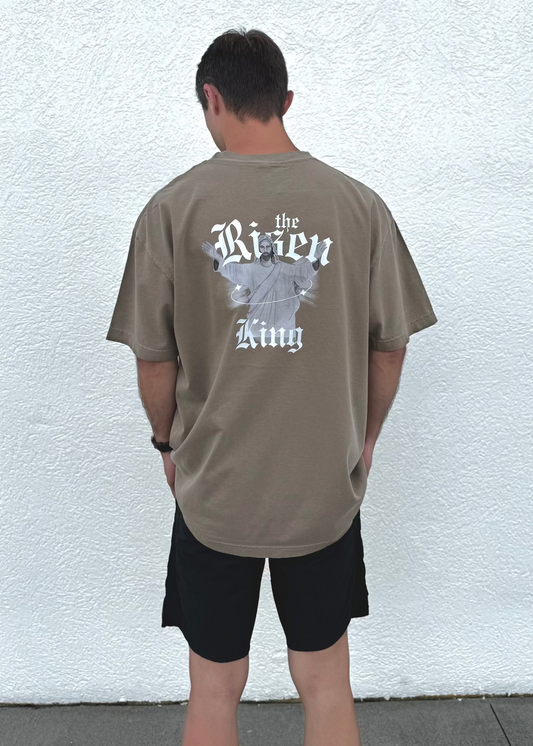 Person wearing a brown t-shirt with text and Jesus graphics on the back, standing against a white wall.