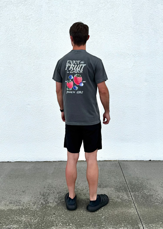 Man wearing a gray t-shirt with fruit design and text, standing against a white wall.