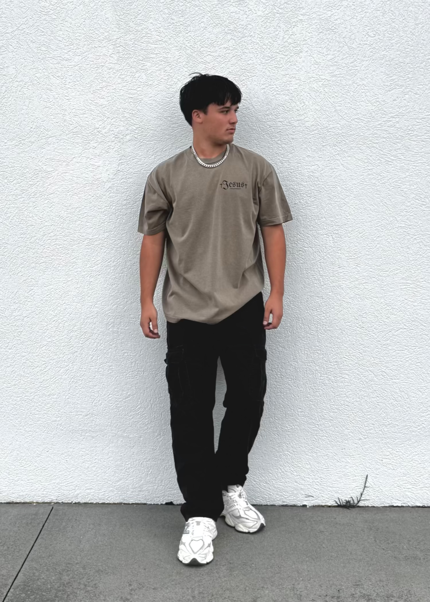 Person wearing a beige t-shirt and black pants standing against a white wall.