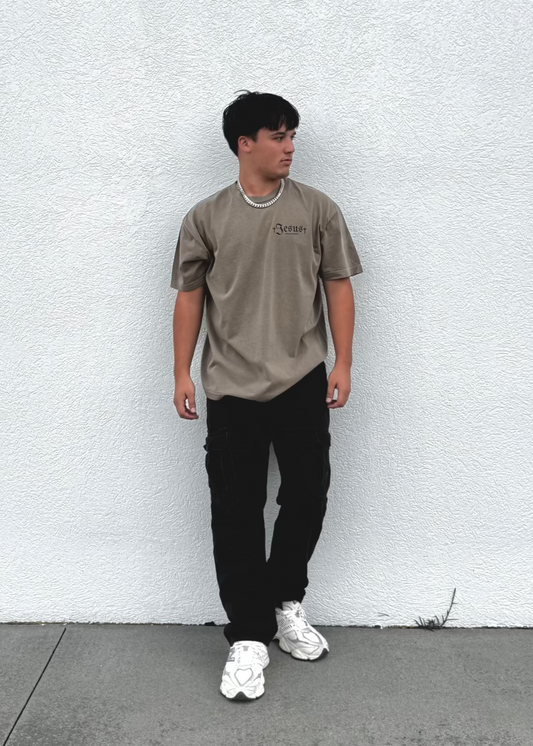 Person wearing a beige t-shirt and black pants standing against a white wall.