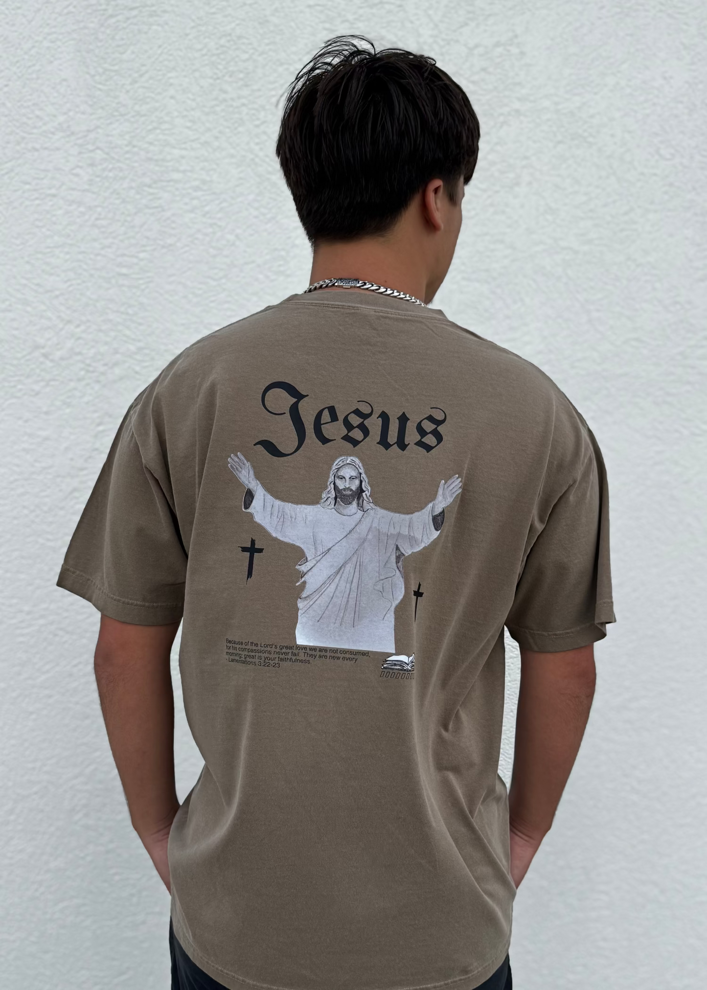 Person wearing a brown t-shirt with 'Jesus' graphic on the back against a white wall.