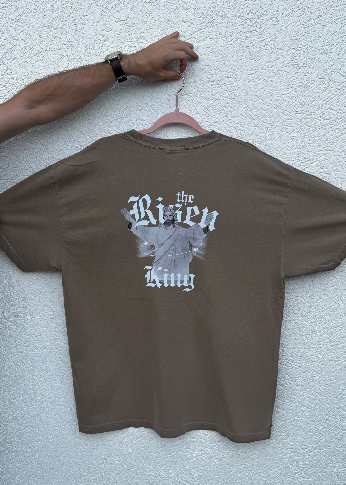 Brown t-shirt with graphic design 'Risen King' on a hanger against a light gray wall