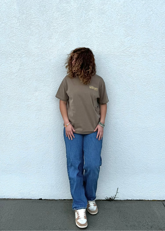 Person wearing a brown t-shirt and blue jeans standing against a light gray wall.