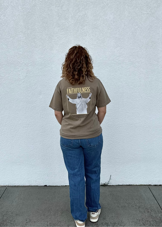 Person wearing a brown t-shirt with a graphic and text, standing against a light gray wall.